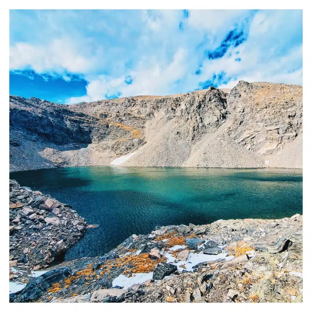 ice blue alpine lake with orange and tan flora around it with tan jagged rocks 