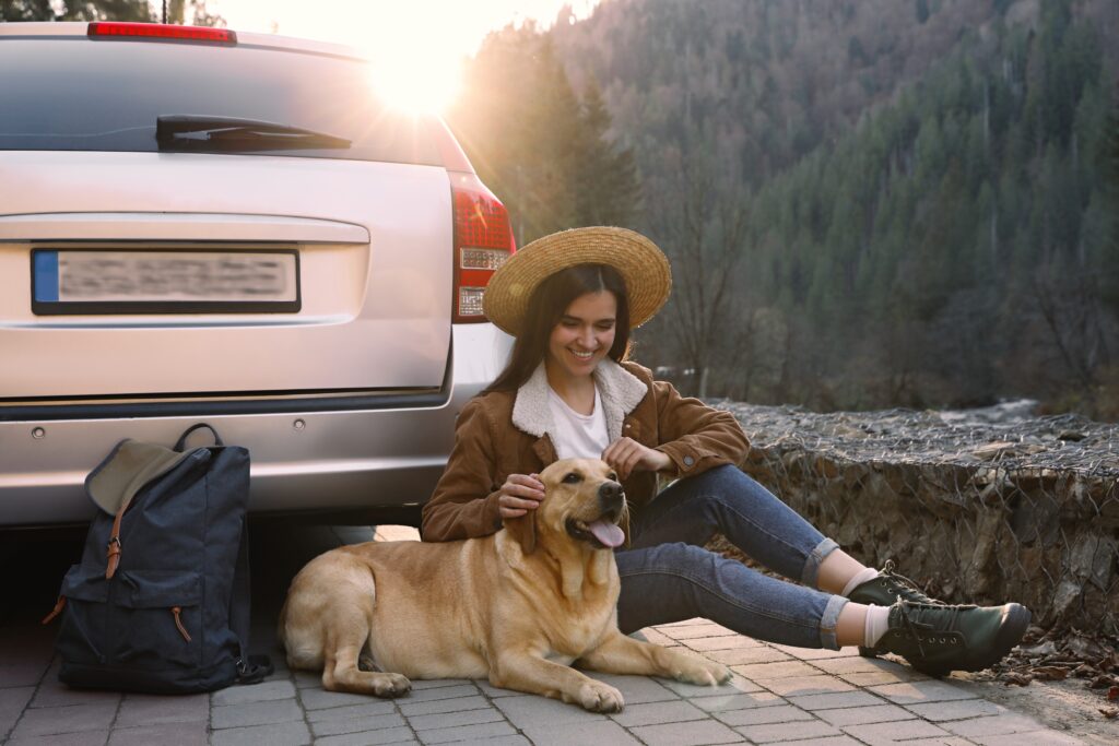 Woman sitting by car with her Labrador dog during a pet-friendly mountain vacation, enjoying the outdoors at sunset with backpack and scenic forest view.